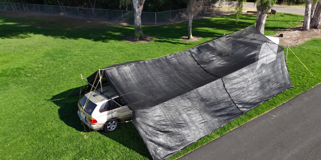 Car parked under a large black shade on a grassy area, hovershade SUV and trailer cabopy setup