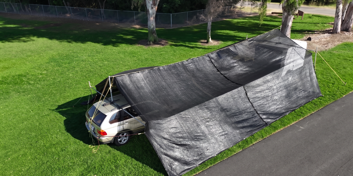 Car parked under a large black shade on a grassy area, hovershade SUV and trailer cabopy setup