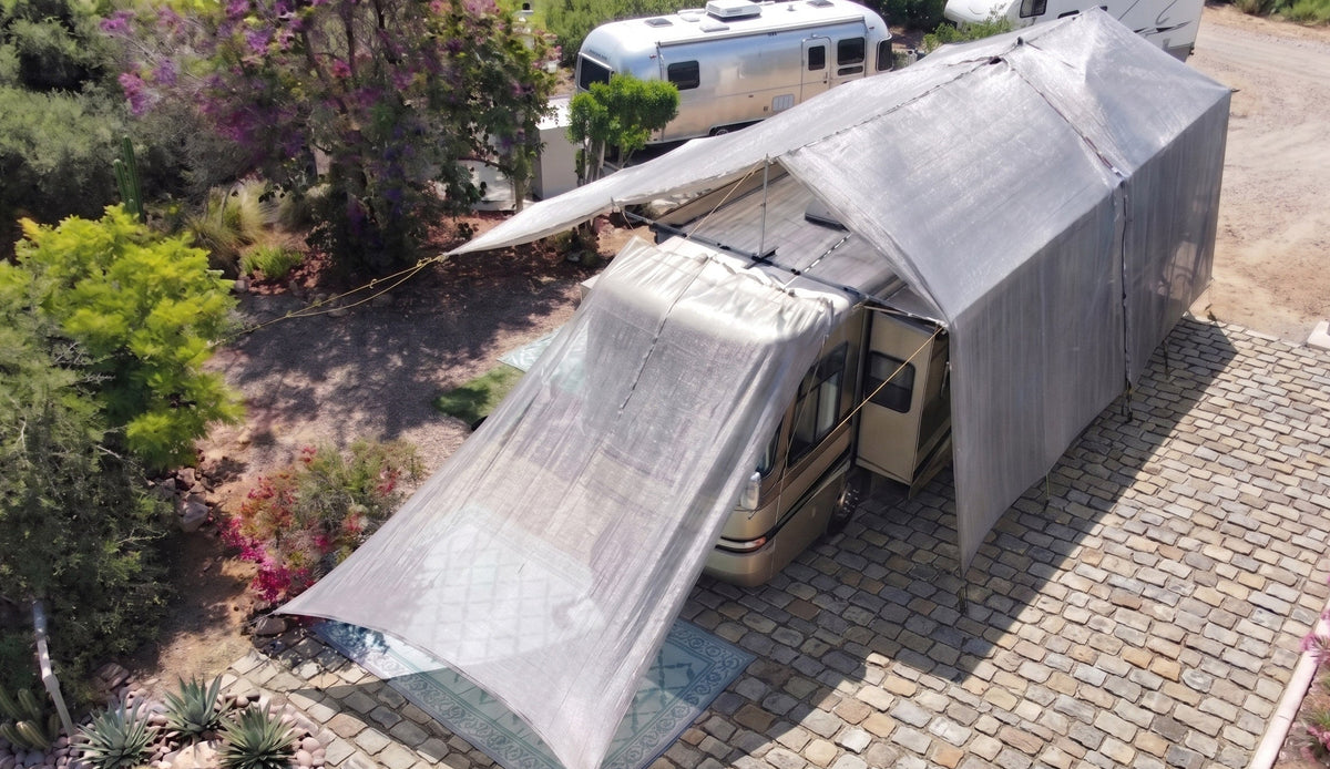 RV with extended hovershade silver shade awning parked on a paved area with trees and flowers in the background