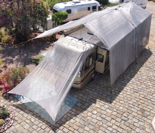 RV with extended hovershade silver shade awning parked on a paved area with trees and flowers in the background